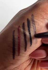 Sex eyelinersviter är ritade på en hand, från djup burgundy till svart. Hudens textur är synlig, och en penna applicerar en ny linje.