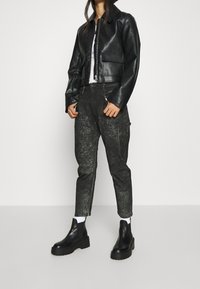 Chaqueta corta de cuero negro con cierre de cremallera y dos bolsillos frontales, combinada con pantalones negros desgastados y botas de tobillo negras gruesas.