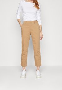 Pantalons en coton beige coupés, avec une coupe droite, deux poches avant et une texture lisse, associés à un haut blanc à manches longues et des baskets blanches.