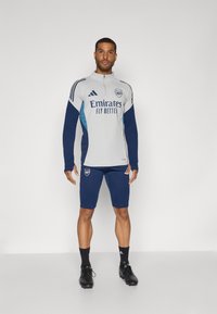 Sportswear featuring a gray zip-up top with blue accents and white logo, paired with navy shorts and black cleats.