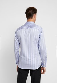 Muž stojící zády, oblečený v světle modré košili s dlouhým rukávem s bílými vertikálními pruhy a černými kalhotami na jednobarevném pozadí.