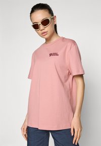 Camiseta em algodão rosa claro com decote redondo, mangas curtas e um pequeno logótipo rosa escuro no lado esquerdo do peito. Corte descontraído, design simples.