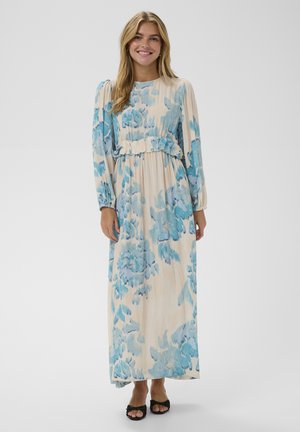 Naine, kes kannab pikkade varrukatega beeži kleiti siniste lillemustritega, mustade avatud varvastega kontsakingadega, seisab ja naeratab ühevärvilise tausta ees.