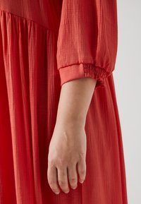 Sarkans teksturēts auduma kleita ar saliektu piedurkni un elastīgu aproci. Izceļ plūstošu dizainu ar smalkiem krokām un mīkstu apdari.