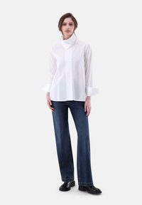 Camicia bianca a maniche lunghe con colletto alto, abbinata a jeans a gamba larga blu scuro e stivali neri alla caviglia. Tessuto fluido, design minimale.