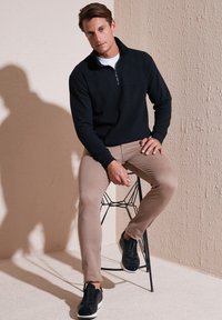 Pullover in pile nero con colletto con zip, pantaloni slim-fit beige e sneakers nere, seduto su uno sgabello in metallo o filo contro un muro testurizzato.