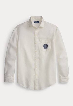 Hvit langermet skjorte med knapper og krage, brystlomme med marineblått brodert skjold, og Polo by Ralph Lauren-merke inne i kragen.