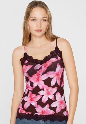 Jeune femme portant un caraco violet foncé avec un imprimé floral rose et une bordure en dentelle noire, debout sur un fond gris clair.