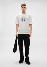 Camiseta blanca de algodón con un gráfico azul de cara sonriente y letras, combinada con pantalones cargo negros y un bolso cruzado negro.