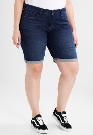 Personne portant un short en denim foncé retroussé, une chemise blanche et des baskets basses noires avec des lacets blancs, debout devant un fond uni.