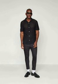 Black short-sleeve shirt with perforated design, button detail, paired with black slim-fit pants and black shoes. Model wears sunglasses and white socks.