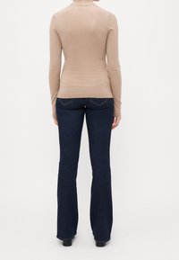 Personne debout, vue de dos, portant un pull à col roulé beige et un jean évasé bleu foncé avec des chaussures noires sur un fond uni.