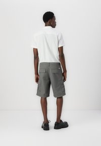 Graue Cargo-Shorts mit seitlichen Taschen, kombiniert mit einem weißen Kurzarmhemd. Schwarze Schuhe, stehend vor einem einfarbigen weißen Hintergrund.