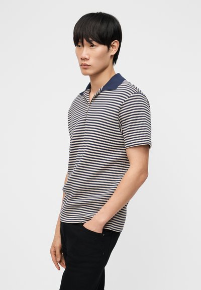 Νέος άνδρας με μαύρα μαλλιά που φοράει μπλε και άσπρη ριγέ πόλο μπλούζα με κοντά μανίκια και μονόχρωμο μπλε γιακά, με τα χέρια στις τσέπες μαύρων παντελονιών.