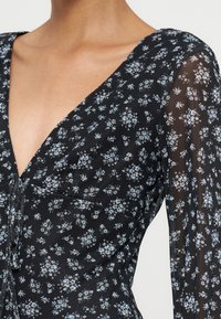 Robe noire avec un petit motif floral blanc et bleu, col en V profond et manches longues transparentes avec le même motif floral.