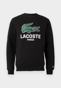 Juodas megztinis, pagamintas iš medvilnės, su žaliu krokodilo logotipu ir baltu tekstu "LACOSTE PARIS". Su gumbuotomis rankovėmis ir apykakle.