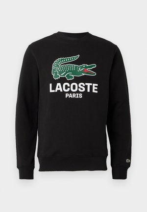 Svart hettegenser i bomull med grønt krokodillemerke og teksten "LACOSTE PARIS" i hvitt. Ribbestrikkede ermer og hals.