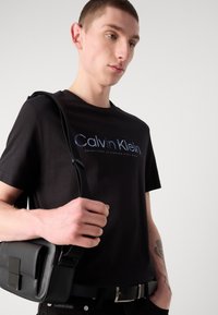 Sort bomulds T-shirt med "Calvin Klein" logo i gradient blå, parret med en sort messenger taske og sort bælte med sølv spænde.