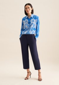 Camicia a maniche lunghe blu a fantasia con bottoni, abbinata a pantaloni eleganti blu navy. Sandali neri con tacco alto e cinturini completano l'outfit.