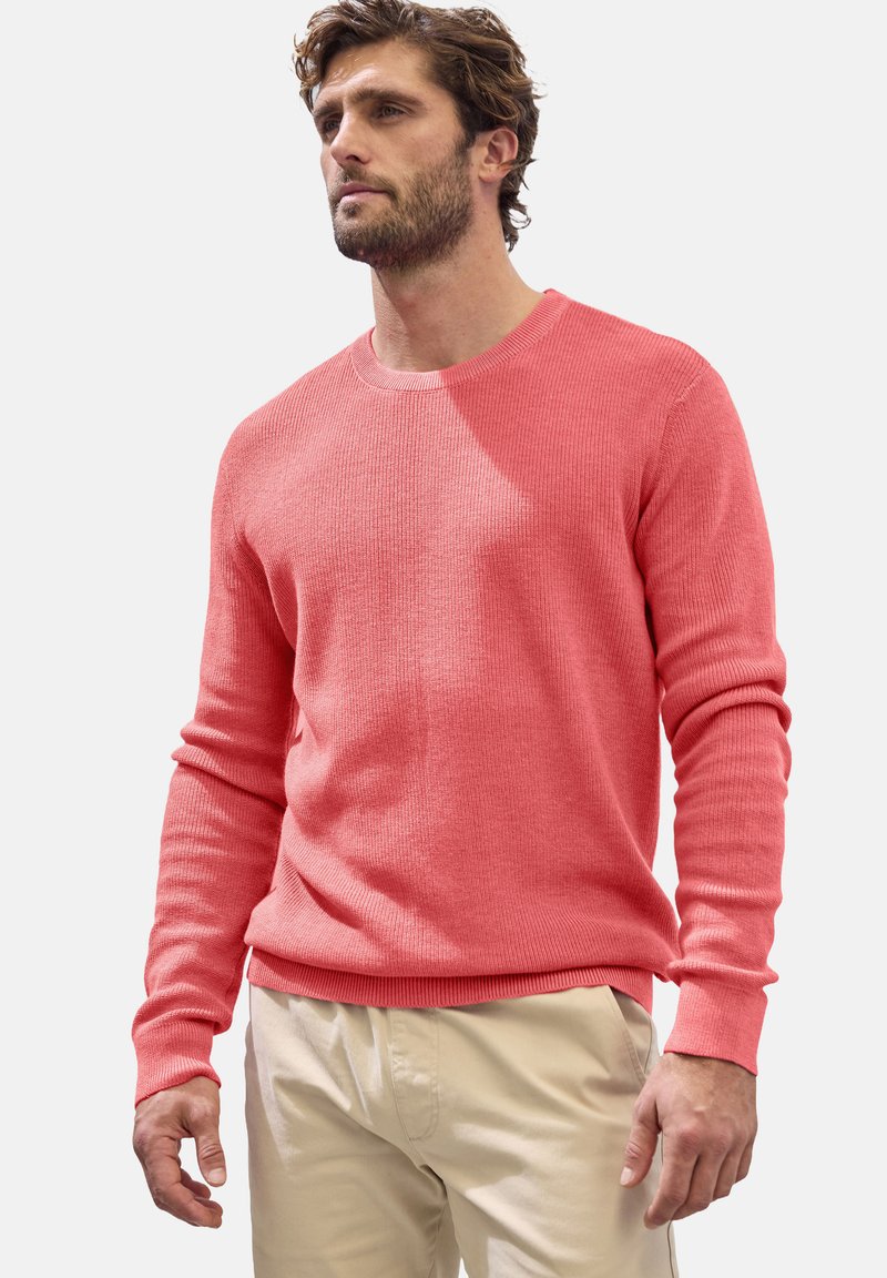 Korallfarbener Strickpullover mit langen Ärmeln und Rundhalsausschnitt. Mit geripptem Textur und Bündchen, getragen über hellbeigen Hosen.