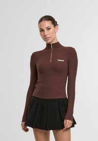 Braunes, geripptes Langarmshirt mit hohem Kragen und halbem Reißverschluss, das ein kleines silbernes Logo auf der Brust trägt. Getragen mit einem schwarzen Plisseerock.