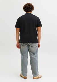 Άνδρας με σγουρά μαλλιά στέκεται γυρισμένος, φοράει μαύρο κοντομάνικο πουκάμισο, ανοιχτόχρωμο τζιν και μπεζ αθλητικά παπούτσια σε απλή φόντο.