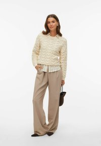 Jersey de punto color crema con un patrón de ondas texturizadas, combinado sobre una camisa de rayas con cuello. Pantalones anchos beige y un bolso oscuro completan el conjunto.