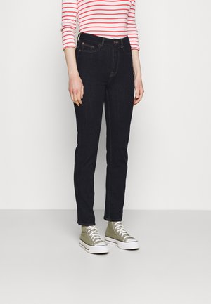 Jeans blu scuro con una vestibilità slim, dotati di cinque tasche e cuciture arancioni, abbinati a sneakers alte verde oliva e a una maglietta a righe a maniche lunghe.