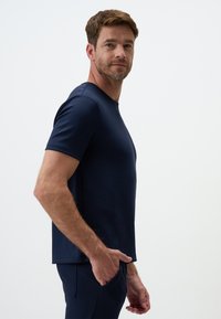 T-shirt blu navy a maniche corte realizzato in tessuto morbido, presenta un colletto rotondo e un taglio dritto. Il modello sta in posa di lato con la mano in tasca.