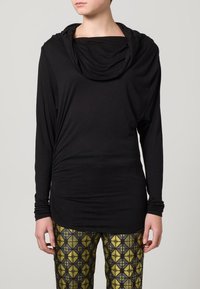 Schwarzes Langarmoberteil mit einem drapierten Rollkragen. Hergestellt aus weichem Stoff, mit strukturierten Details und enganliegenden Ärmeln, kombiniert mit geometrisch gemusterten Leggings.