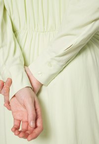 Mains jointes dans le dos, portant une robe verte pâle à manches longues avec des poignets boutonnés et une taille froncée.