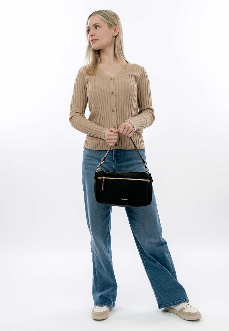 Black leather handbag with a zippered pocket, held by a person in a beige ribbed cardigan and blue jeans, standing against a white background.