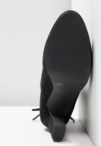Bottine en daim noir avec un talon effilé et une semelle en caoutchouc texturée. Indicateur de taille 37 embossé en or sur le côté de la semelle.