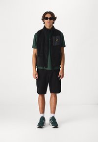 Black fleece vest with a zip, green t-shirt, black cargo shorts, white socks, and dark green sneakers. Accessories include black sunglasses.