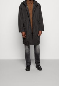 Gabán impermeable negro con capucha, bolsillos con cremallera y un corte relajado, combinado con una camisa de botones marrón y jeans grises.