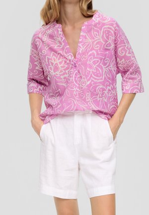 Woman wearing a pink patterned button-up blouse with rolled sleeves and white high-waisted shorts, hands in pockets, standing.