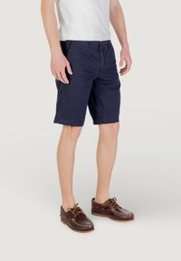 Shorts in cotone blu navy con chiusura a bottoni, tasche laterali e orli cuciti, abbinati a scarpe da barca in pelle marrone con dettagli in pizzo.