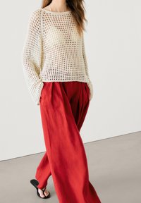 Pull tricot beige clair avec un motif en maille ouverte, associé à un pantalon ample, taille haute et rouge vif. Des sandales plates complètent le look.