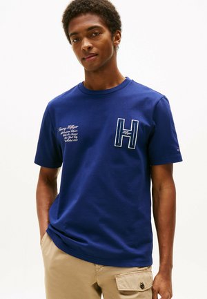 Camiseta de manga corta azul marino de algodón con cuello redondo. Presenta la letra "H" en costura azul claro y texto de la marca impreso en el frente.