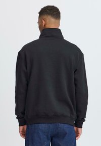 Sweat-shirt noir avec un col montant et une coupe décontractée, fabriqué dans un tissu doux. Présente des poignets et un ourlet côtelés, associé à un jean en denim bleu.