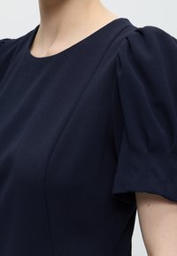 Gros plan d'une personne portant une robe bleu marine avec des manches courtes bouffantes et un col rond.