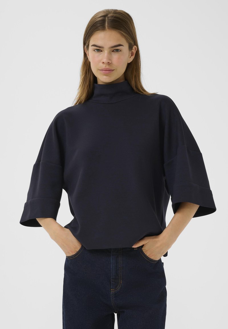 Námorně modrý oversize top s vysokým výstřihem a širokými rukávy. Měkká, hladká tkanina. Modelka ho nosí s tmavými džíny, s rukama v kapsách.