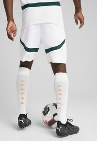 Shorts de sport blancs avec des accents verts et des motifs texturés. Chaussures de football noires avec une finition texturée ; chaussettes blanches avec des détails en forme de flèche orange.