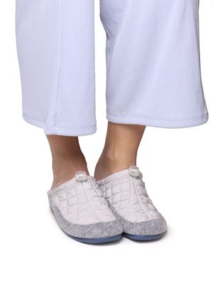 Chaussons gris matelassés avec une partie supérieure blanche, présentant une pointe arrondie et un accent circulaire près de l'ouverture, posés sur un pantalon de couleur claire.