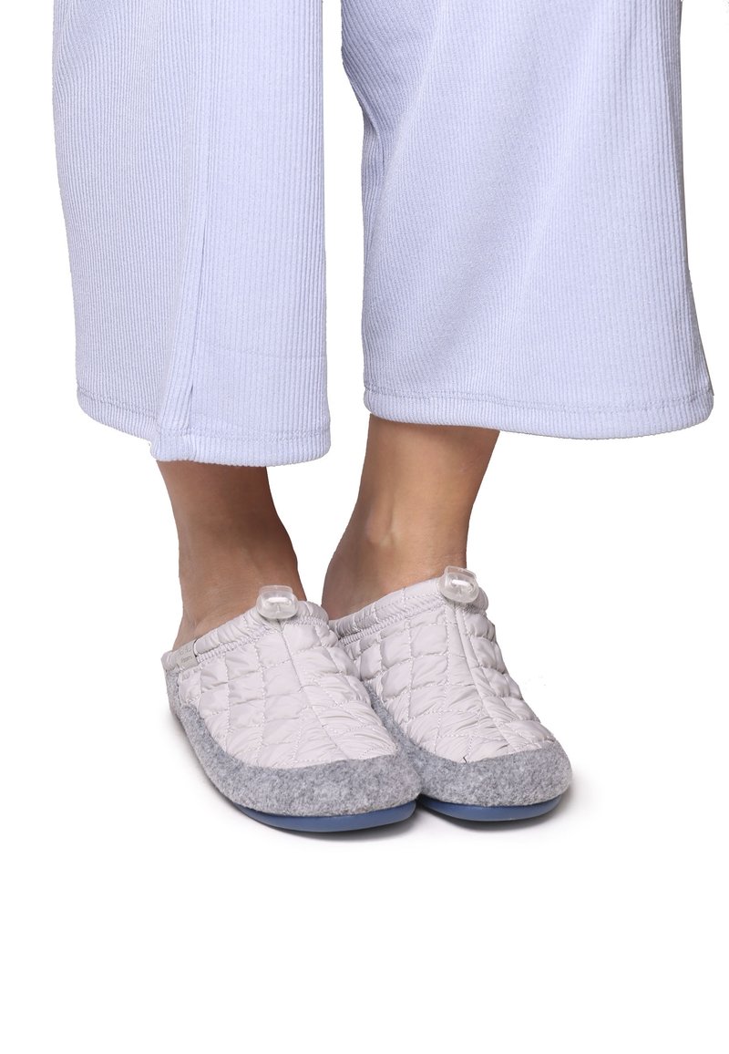 Chaussons gris matelassés avec une partie supérieure blanche, présentant une pointe arrondie et un accent circulaire près de l'ouverture, posés sur un pantalon de couleur claire.