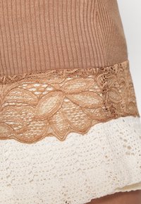 Brun ribbad tyg upptill, prydd med spetstrimma med blommönster i en ljusare tan, lager på lager över vit virkat nertill.