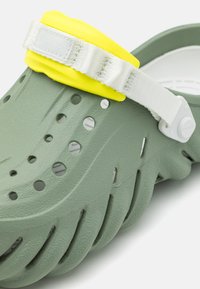Green rubber clog featuring a textured surface, multiple ventilation holes, and a detachable yellow strap with white hardware.