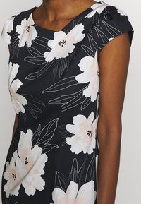 Mujer con un vestido negro con grandes estampados de flores blancas y rosa pálido y mangas cortas, mostrada desde el cuello hasta la mitad del torso.