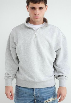 Jonge man met een lichtgrijs halfgezipte trui en gescheurde blauwe spijkerbroek, staand tegen een effen witte achtergrond.