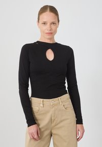 Svart langermet topp med nøkkelhullsutskjæring i halsen, laget av glatt stoff, paret med lysebeige høyt midjebyxor.